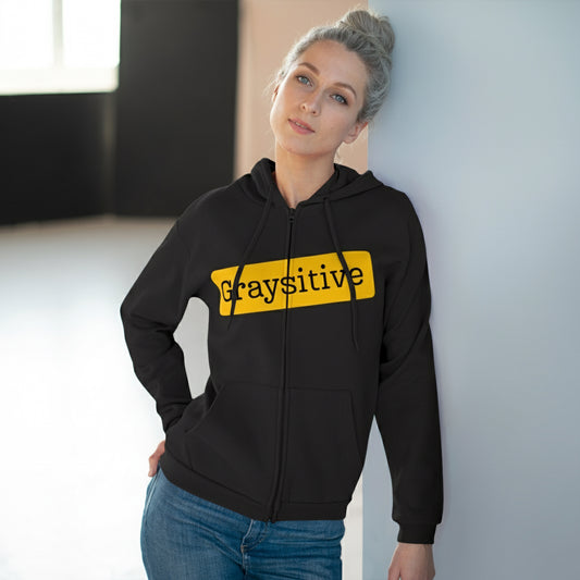 Person wearing a black hoodie with 'Graysitive' text in yellow, standing against a light-colored wall.